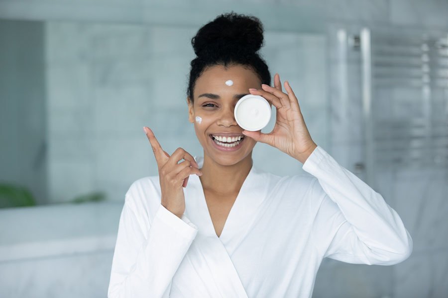 Heureuse belle jeune femme multiraciale d'ethnie africaine en peignoir tenant un pot de crème, satisfaite des résultats des produits cosmétiques professionnels, routine de soin quotidienne efficace dans la salle de bain.