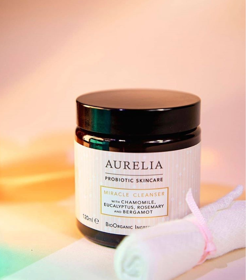 Des nuances de lumière arc-en-ciel entourent une bouteille de nettoyant miracle Aurelia, indispensable pour ces tendances beauté estivales. Une mousseline de bambou rose se trouve à côté de la bouteille.