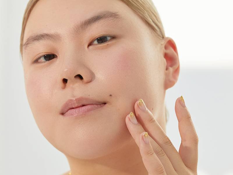 Person with blonde hair pulled back looking at the camera and touching their fingers to their jaw