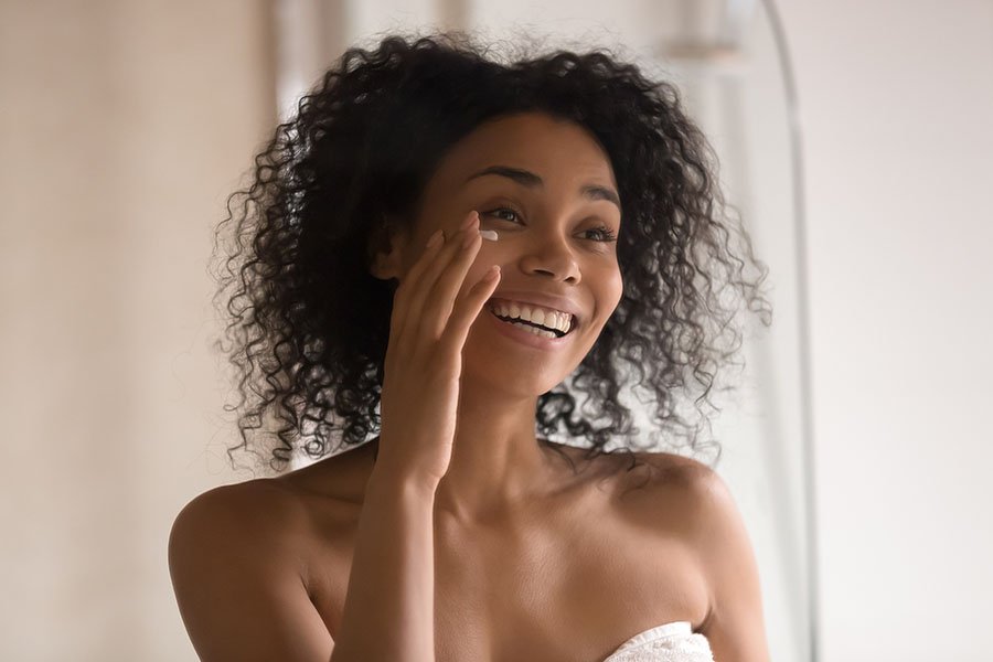 Souriante jeune femme afro-américaine enveloppée dans une serviette après la douche, regarde dans le miroir, applique une crème anti-âge sous les yeux, heureuse femme biraciale effectue des procédures de beauté de soins de la peau le matin à la maison